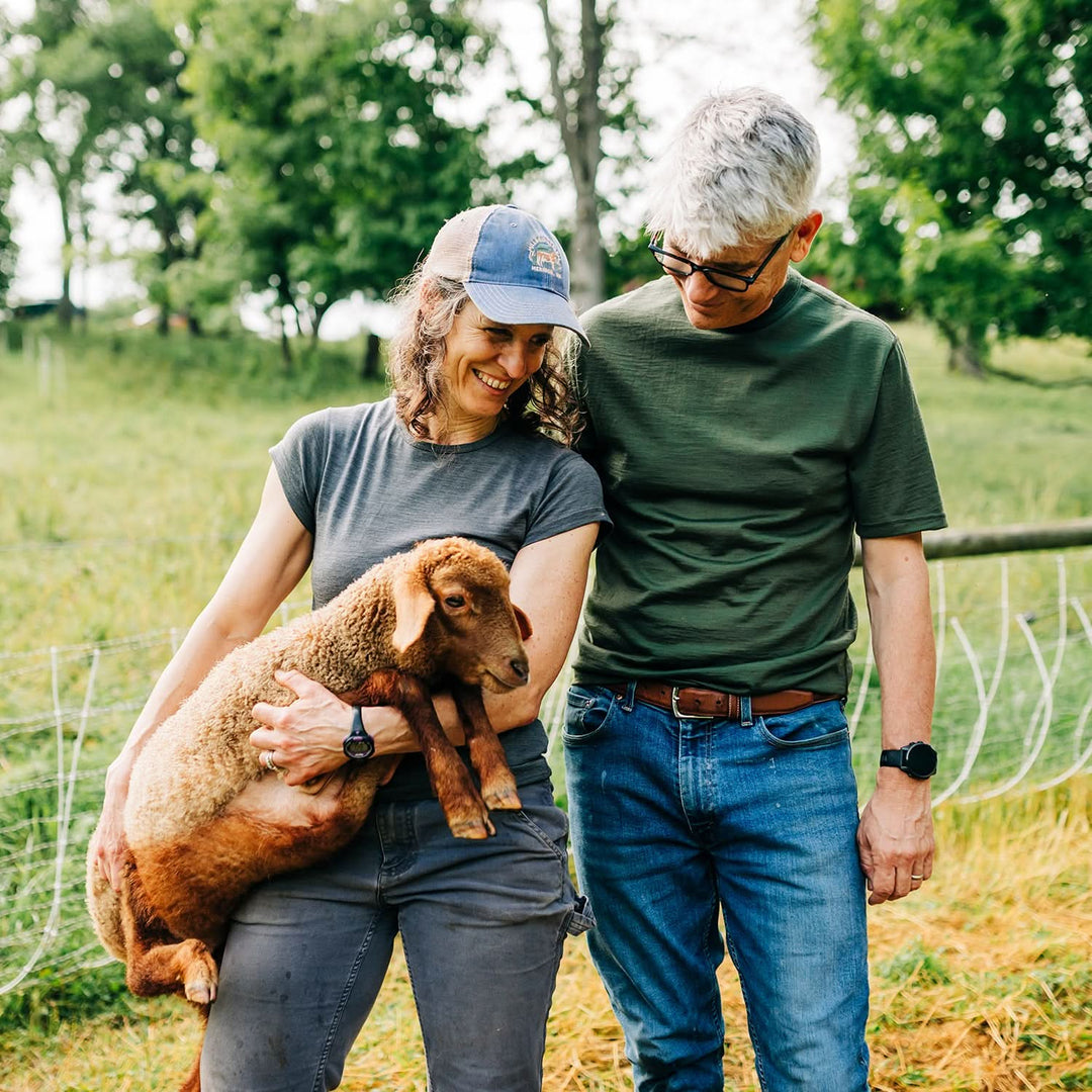 Two people standing in a field with a small lamb, surrounded by trees and greenery. in lightweight merino wool tee shirts 