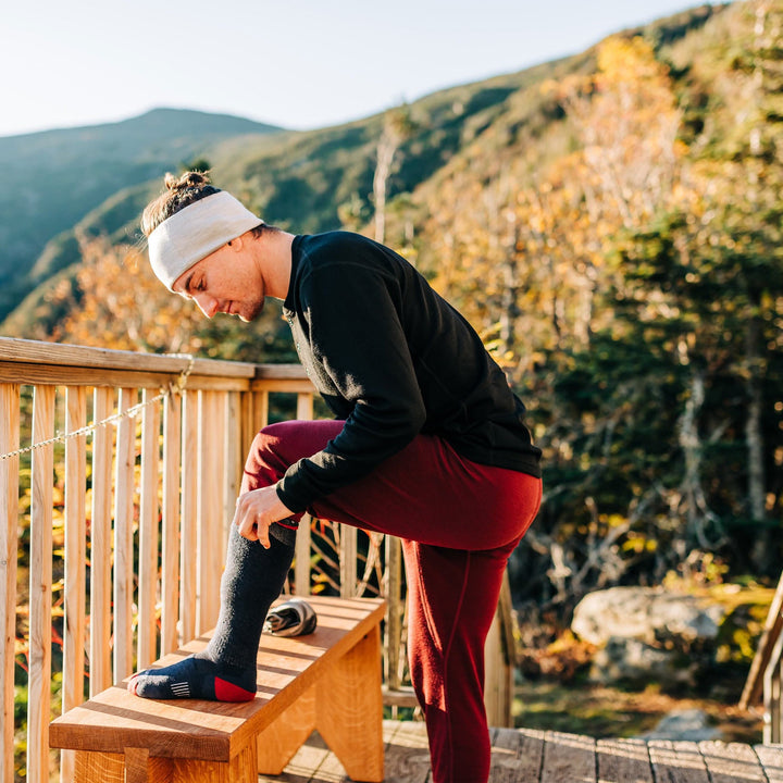 Lightweight over the calf merino wool socks. Made in USA. All season, woman pulling up a sock on a mountain lodge deck 