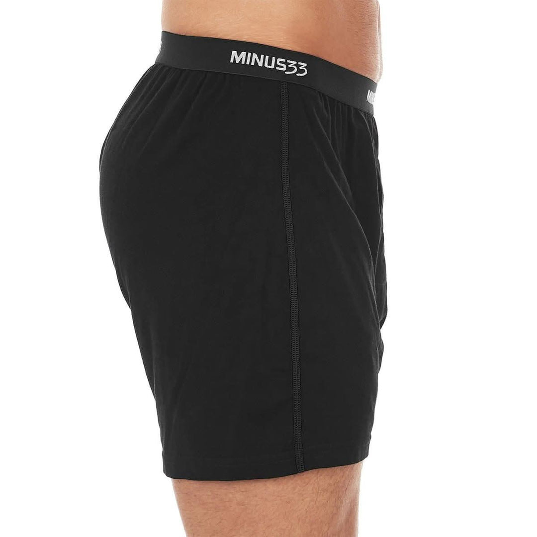 Close-up front view from waist to mid-thigh of a man wearing black Woolverino micro weight merino wool boxer shorts, showing the elastic waistband, button fly, relaxed fabric, and loose leg openings 