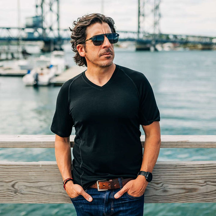 Man wearing sunglasses and a black merino wool t-shirt by a waterfront with boats and a bridge in the background. 