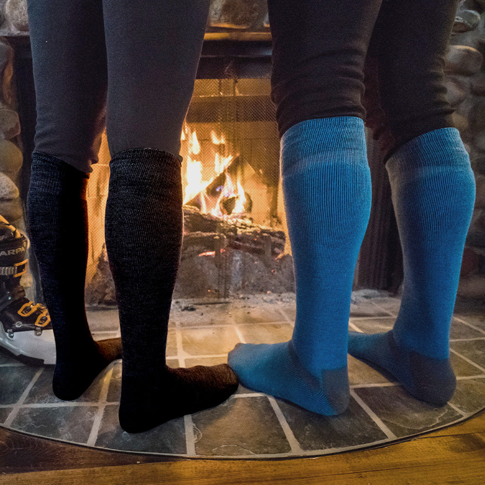 Two pairs of legs wearing black and blue merino wool ski socks standing in front of a fireplace.