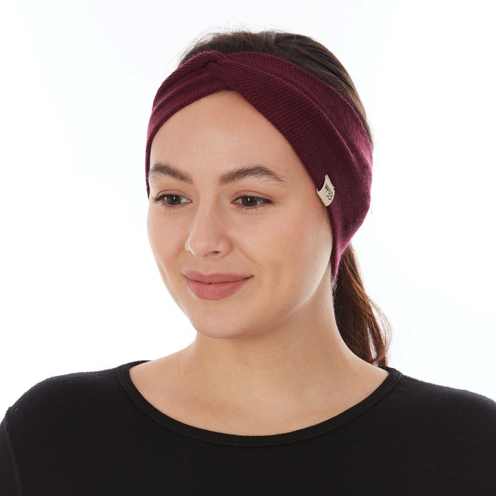 Three-quarter front view of a woman wearing a burgundy midweight merino wool knit twist headband, showing the crossed center front, wide ear coverage, and small Minus33 tag at the right temple 