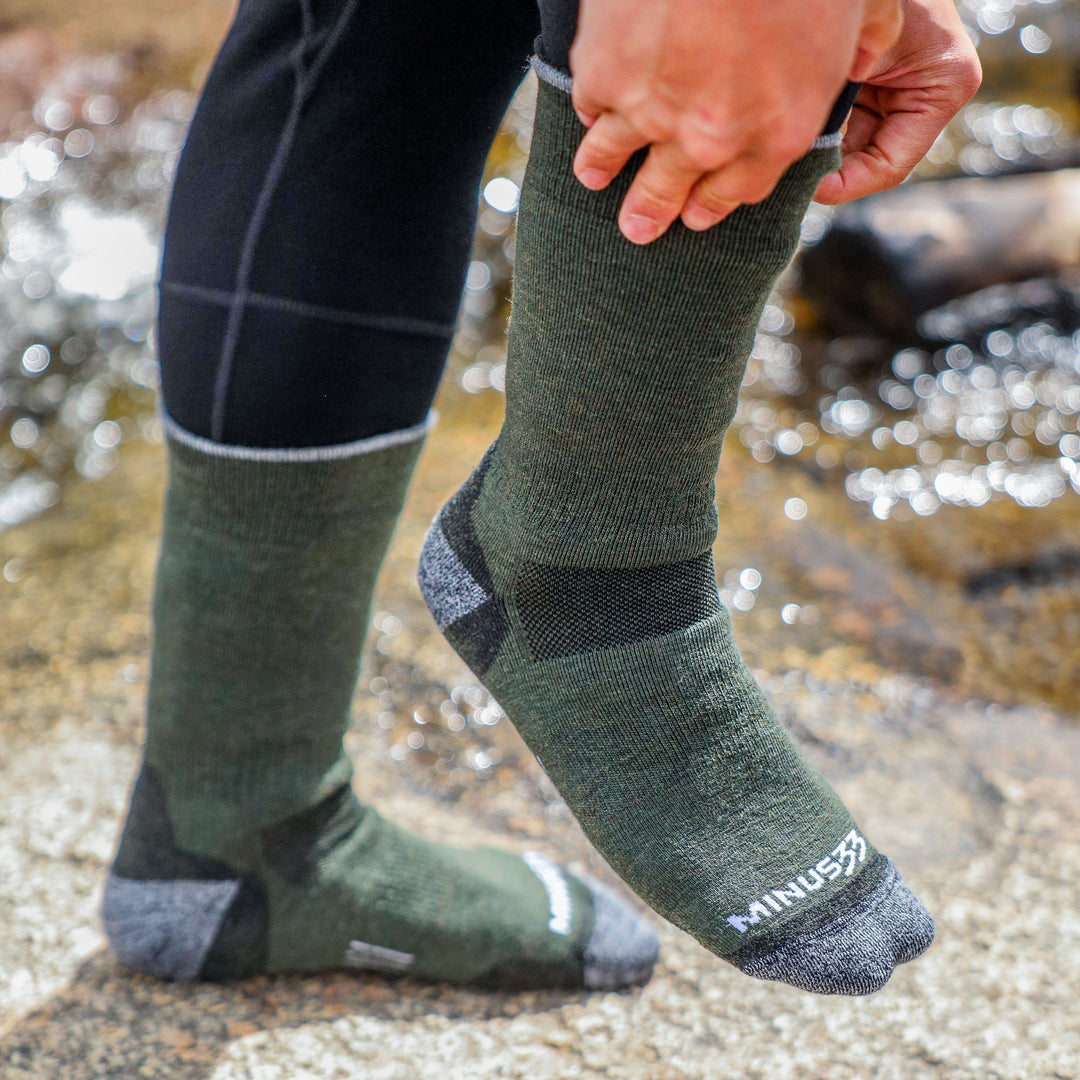 Close-up side view of a person pulling on an olive green midweight merino wool crew hunting sock over black leggings beside a stream, showing the gray reinforced toe and heel and white Minus33 logo 