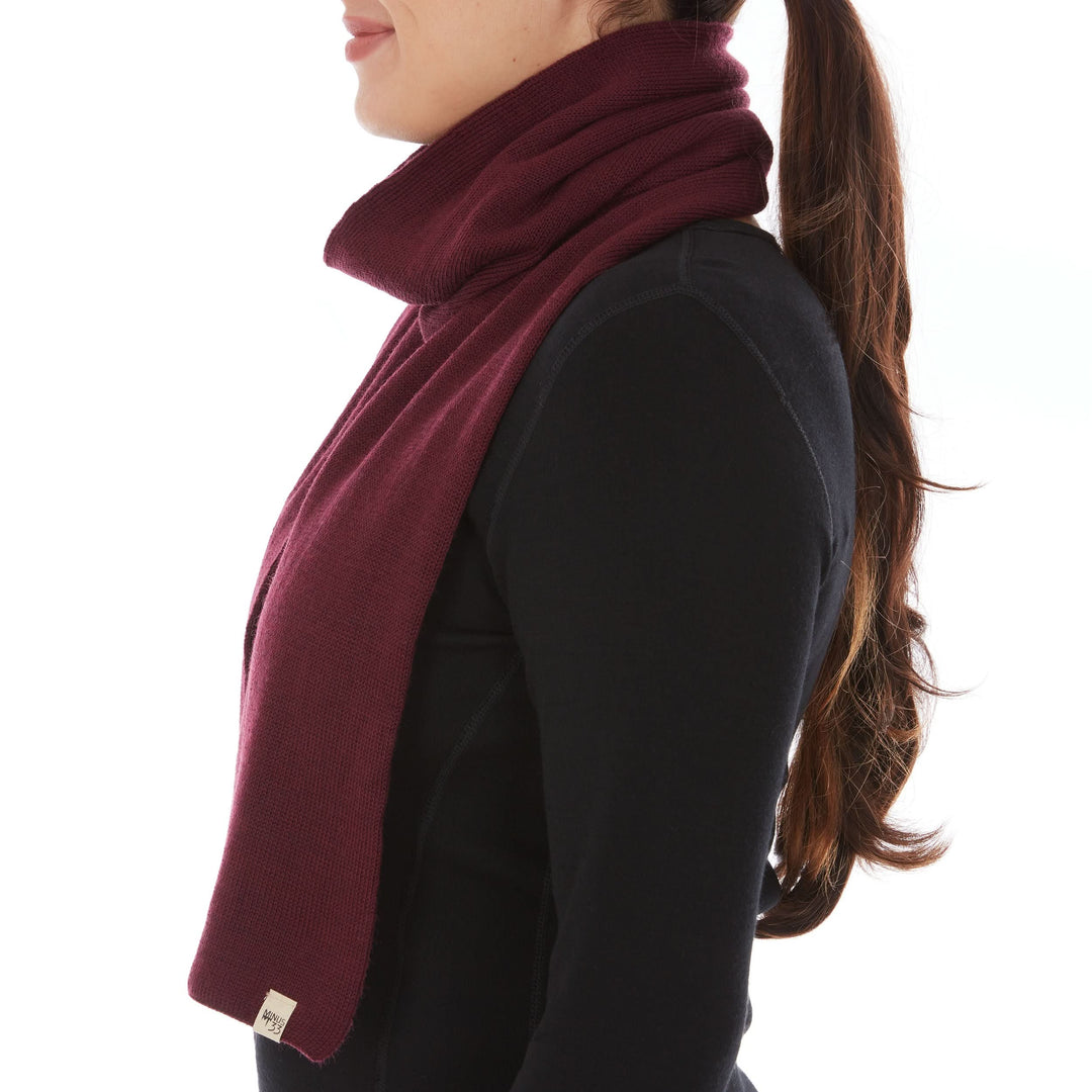 Left-facing side view of a woman wearing a burgundy midweight merino wool knit scarf loosely wrapped around her neck, with one long end hanging down the front of a black top and a Minus33 tag near the tip 