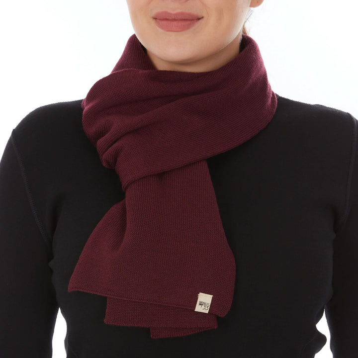 Front close-up view of a woman wearing a burgundy midweight merino wool knit scarf wrapped twice around her neck, with two broad ends layered over a black top and a small Minus33 tag near the lower edge 
