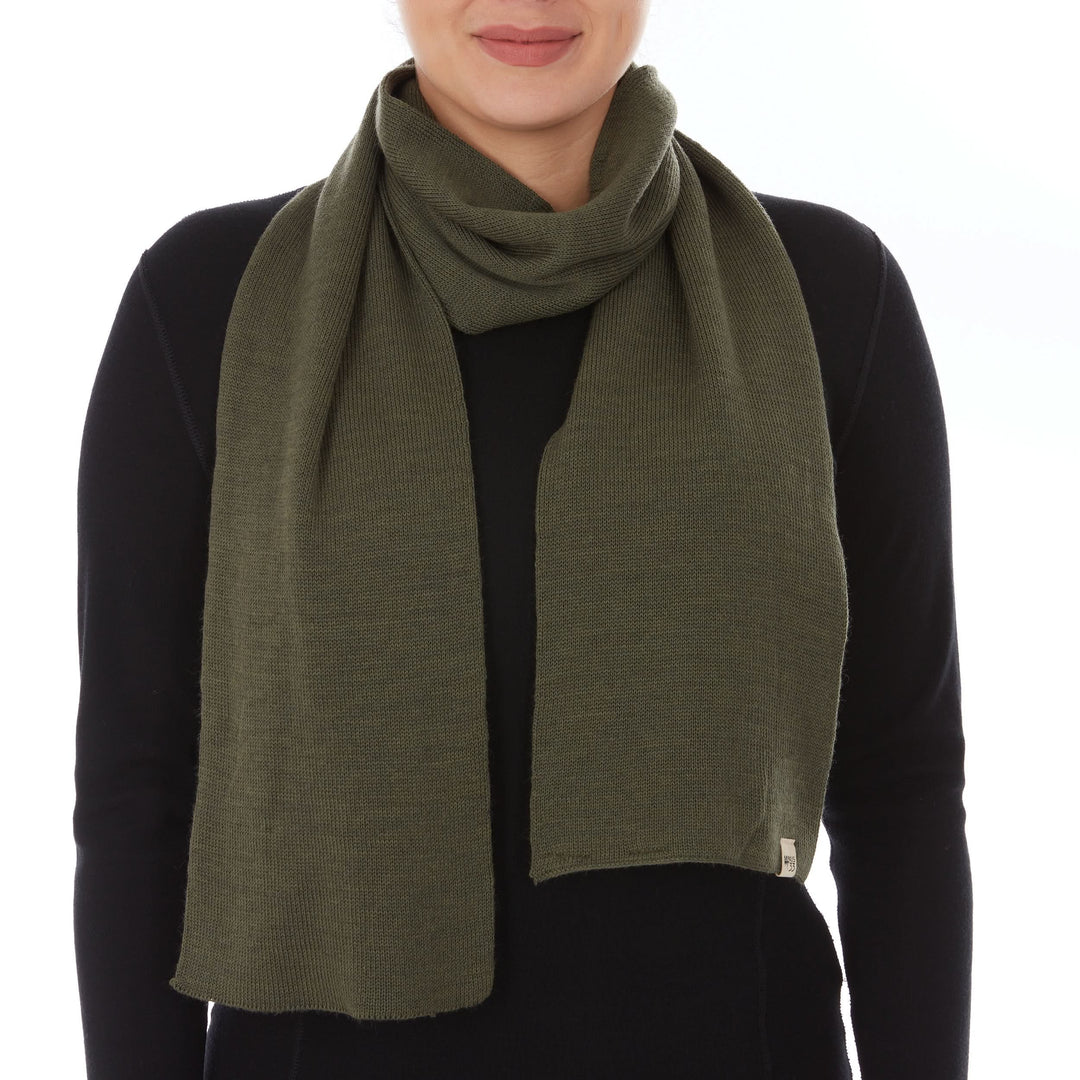 Front view of a woman wearing an olive drab green midweight merino wool knit scarf looped once around her neck, with two wide uneven ends hanging over a black long-sleeve top and a small Minus33 tag at the lower right 