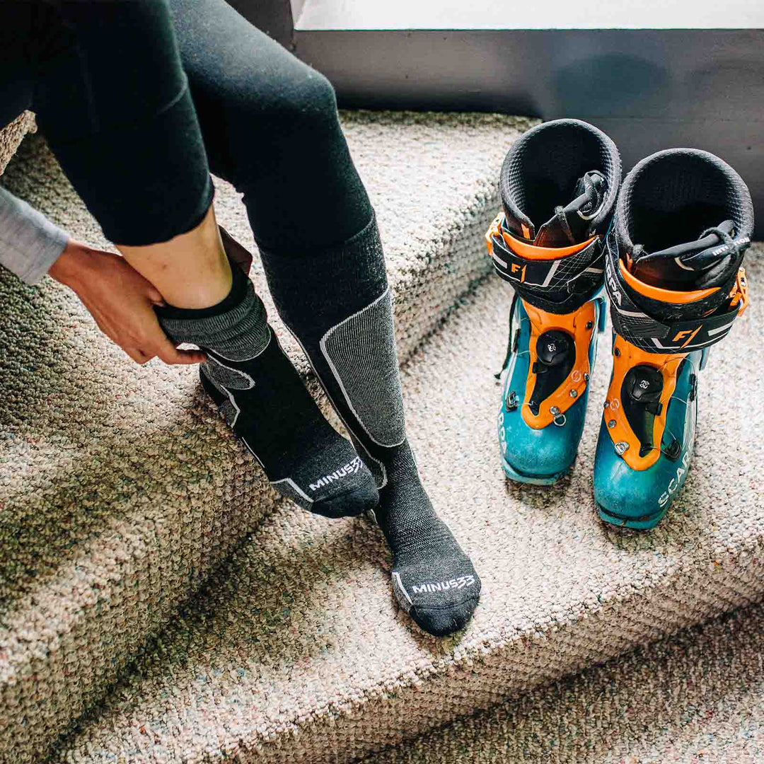 Right side view of a black micro weight merino wool light-cushion over-the-calf snowboard sock being worn, showing the tall ribbed cuff, padded shin, shaped heel, and reinforced toe 