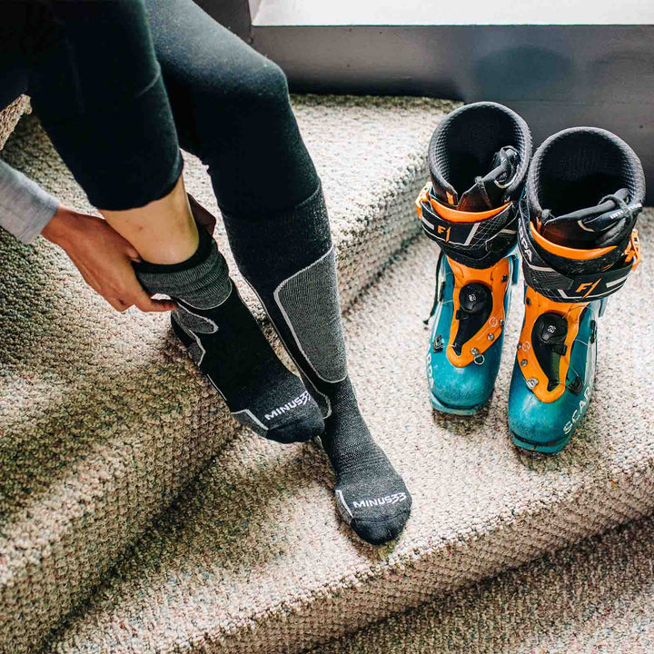 Right side view of a black lightweight merino wool cushioned over-the-calf snowboard sock being worn, showing the tall cuff, padded shin, contoured ankle, shaped heel, and reinforced toe 