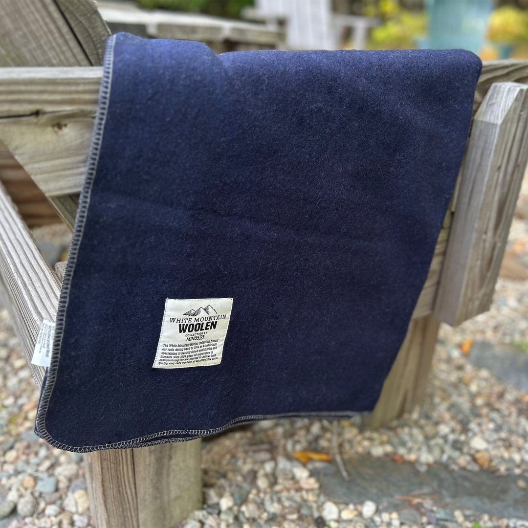 Close-up front view of a solid navy lodge wool blanket draped over the arm and back of a weathered wooden outdoor chair, with contrasting stitched binding and the White Mountain Woolen Mill label visible 