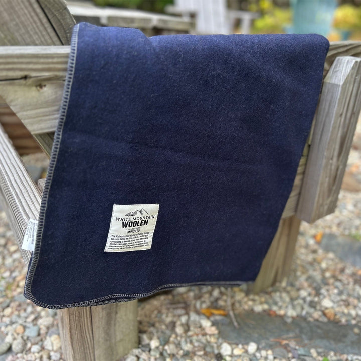 Close-up front view of a solid navy lodge wool blanket draped over the arm and back of a weathered wooden outdoor chair, with contrasting stitched binding and the White Mountain Woolen Mill label visible 