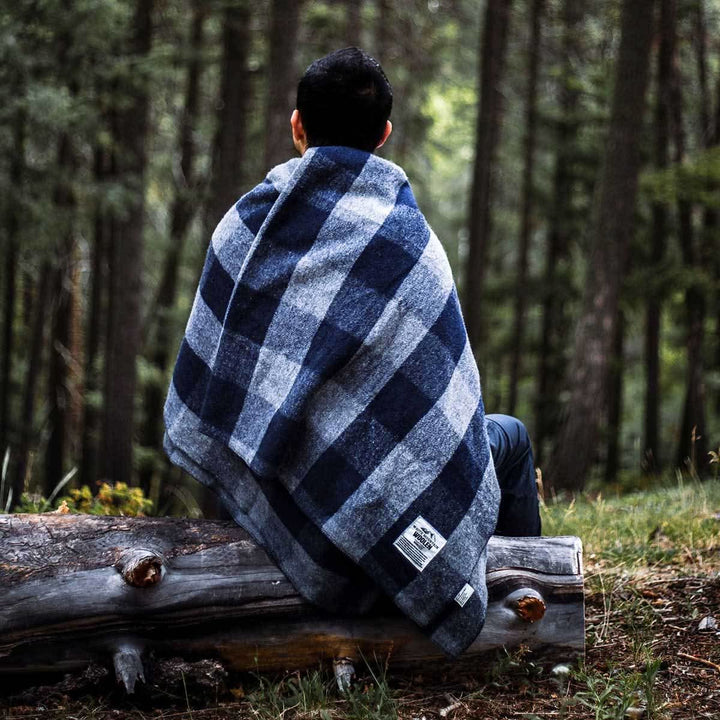 Rear view of a man seated on a fallen log in a pine forest, wrapped around the shoulders in a gray-and-blue buffalo plaid lodge wool blanket with the brand label visible near the hem 