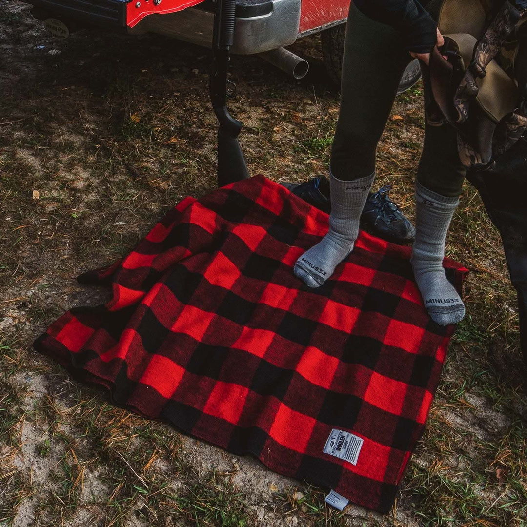 Downward front view of a person wearing gray crew socks while standing on a red-and-black buffalo plaid wool blanket beside a truck, holding boots and preparing to put them on 