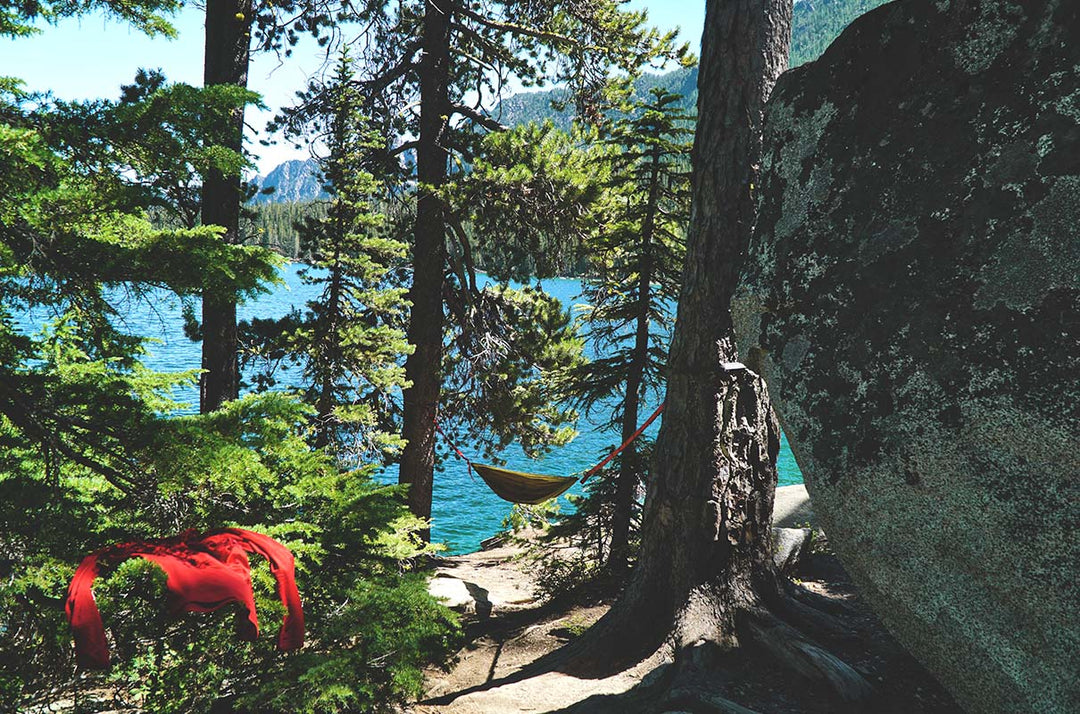 Wide outdoor view of a red merino wool long-sleeve base layer shirt air-drying over an evergreen branch beside a lakeside hammock, pine trees, and a large granite boulder