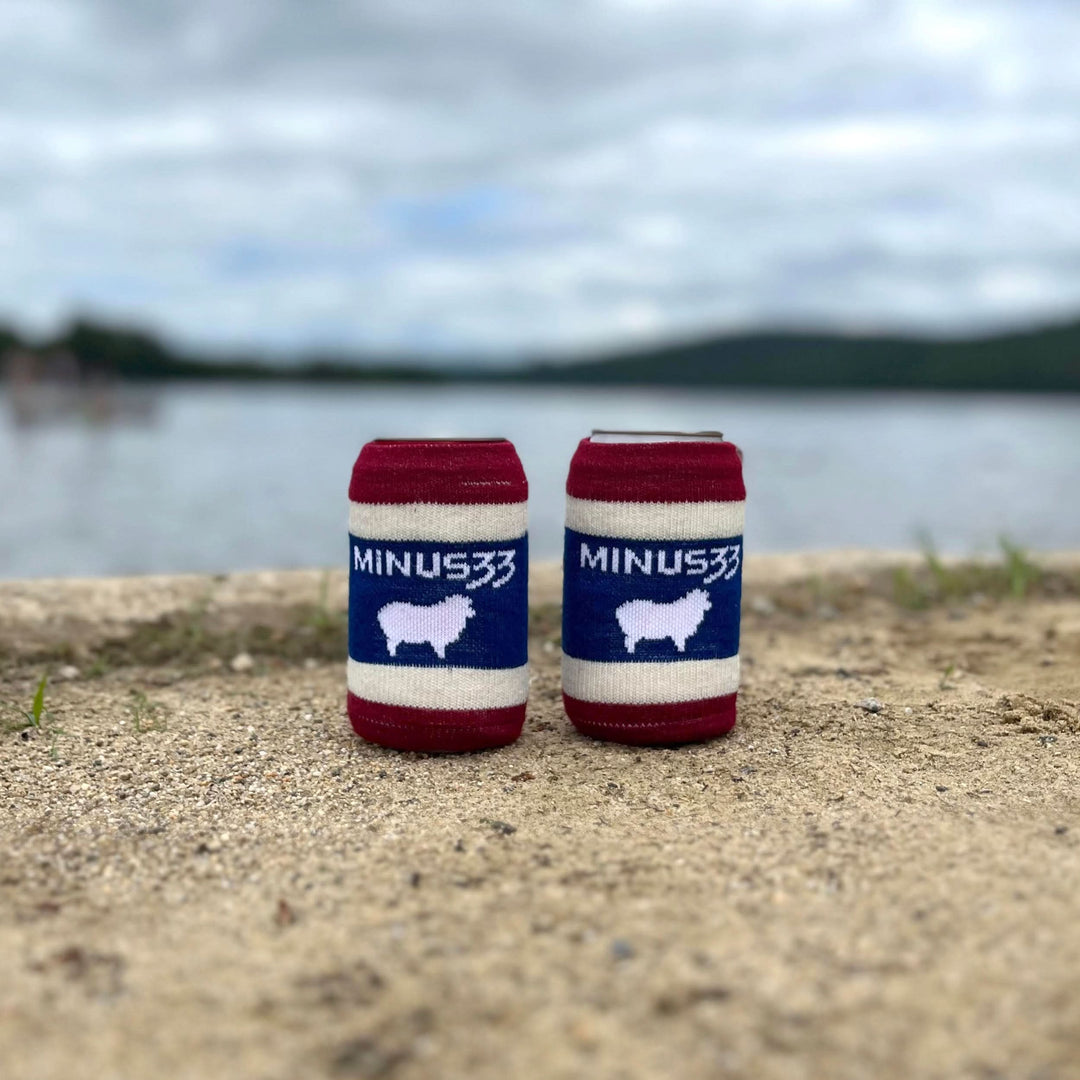 Front view of two red, white, and navy knit Minus33 Merino Cool-Zie can sleeves standing on a sandy lakeshore, each displaying the Minus33 name and a white sheep graphic against a blurred lake and hills