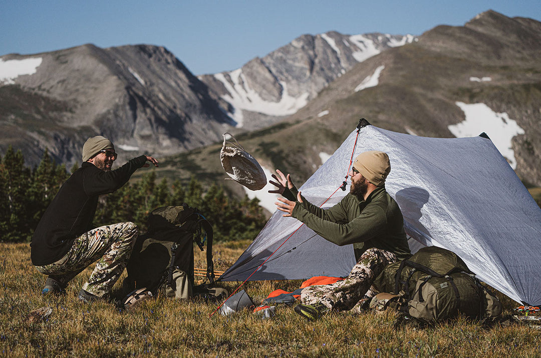 Camping scene with two men wearing layered merino wool hunting clothing beside a campsite, showing long-sleeve tops, rugged outdoor pants, trees, and camping equipment in the background