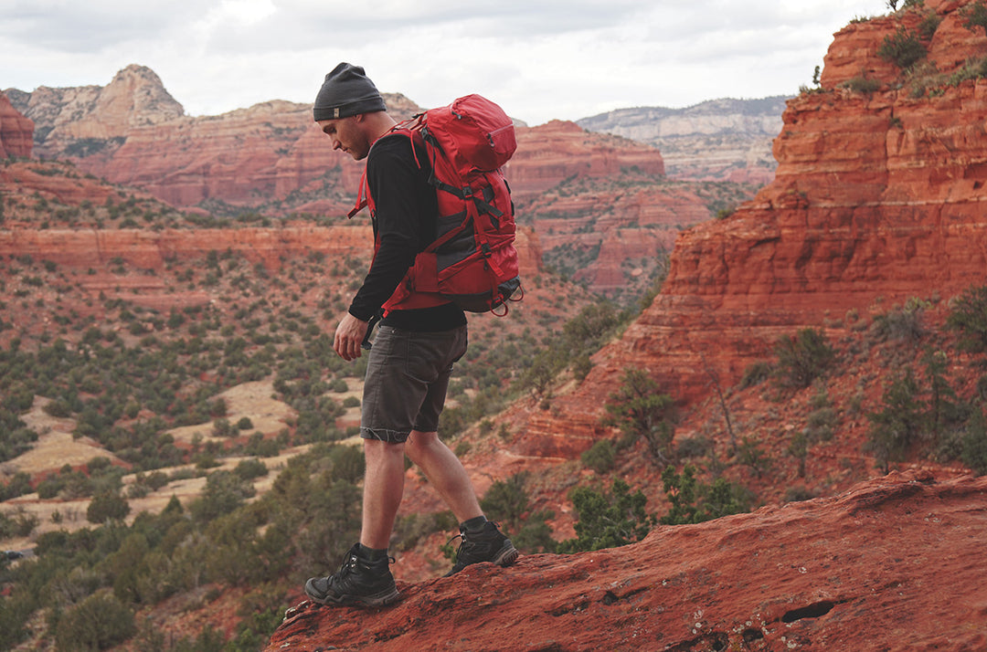 Full side view of a man wearing a black merino wool long-sleeve shirt, dark beanie, gray hiking shorts, and a large red backpack as he walks along a red-rock canyon ridge in Arizona