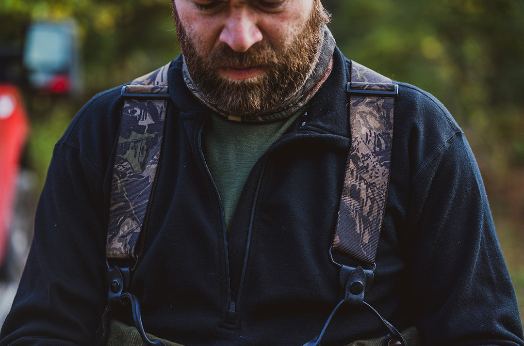Close-up front view of a bearded hunter wearing a black merino wool Expedition quarter-zip midlayer over a green base layer, with camouflage suspender straps crossing his shoulders