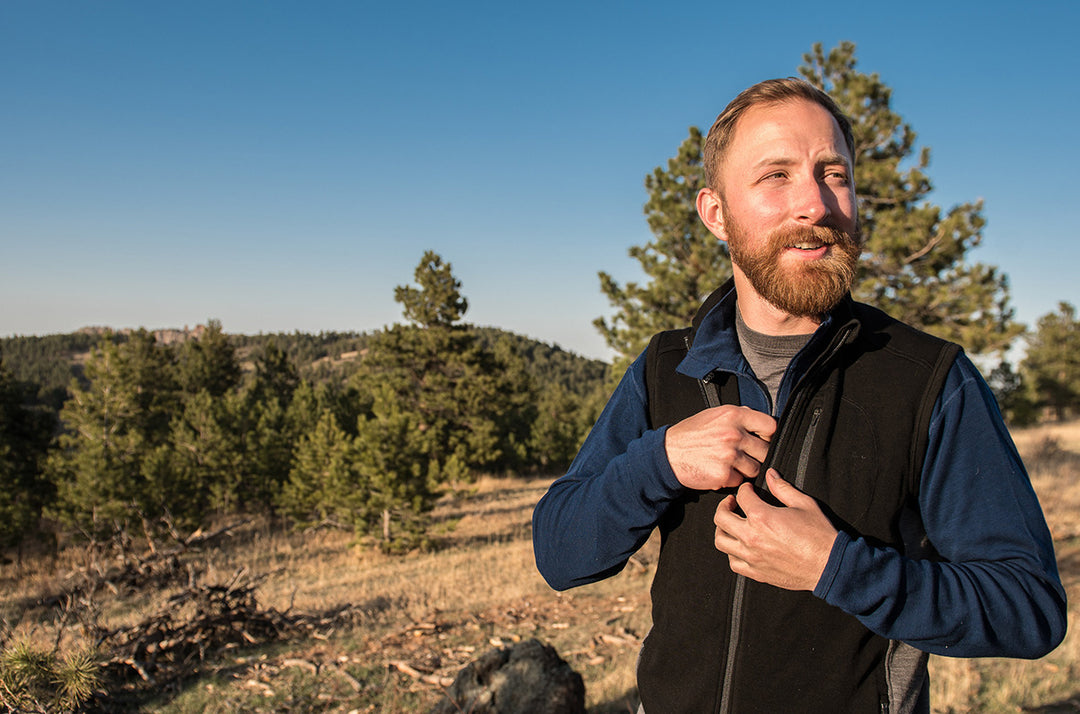 Front three-quarter outdoor view of a bearded man zipping a black merino wool insulating vest over a navy long-sleeve base layer while standing on a sunlit pine-covered hillside