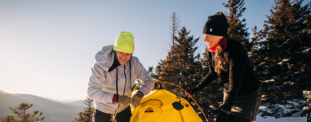 Wide front view of a man and woman wearing hoodies and merino wool beanies while setting up a yellow tent on a snowy mountain