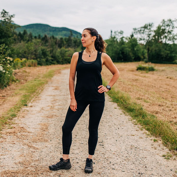 Side view of a woman wearing a black micro weight merino wool racerback tank top, showing the scoop neckline, narrow strap, deep armhole, fitted waist, and hip-length hem 