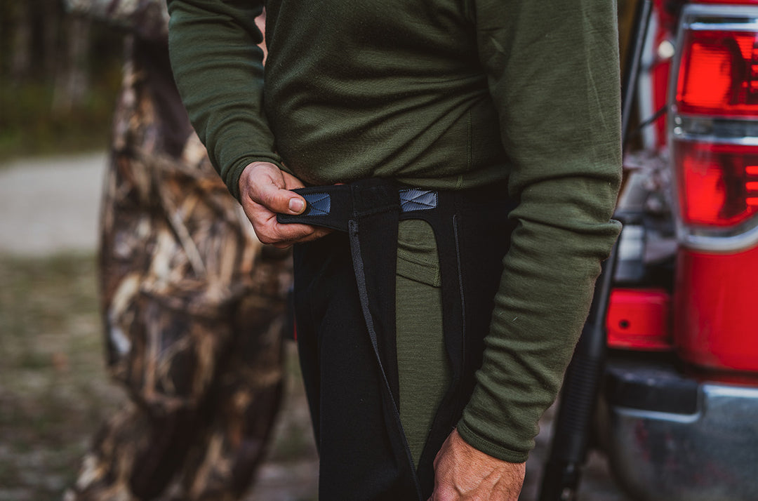 Close-up front view of a hunter fastening the waistband of black zip-front merino wool midlayer pants over an olive green long-sleeve base layer beside a red pickup truck