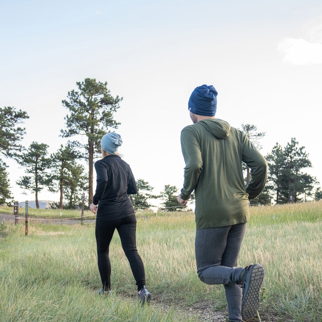 Back action view of a man trail running through a grassy pine clearing in gray midweight merino wool flyless base-layer bottoms, an olive hooded top, and navy beanie, following a woman dressed in black with a gray beanie