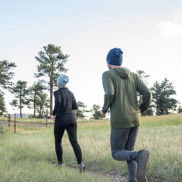 Back action view of a man trail running through a grassy pine clearing in gray midweight merino wool flyless base-layer bottoms, an olive hooded top, and navy beanie, following a woman dressed in black with a gray beanie
