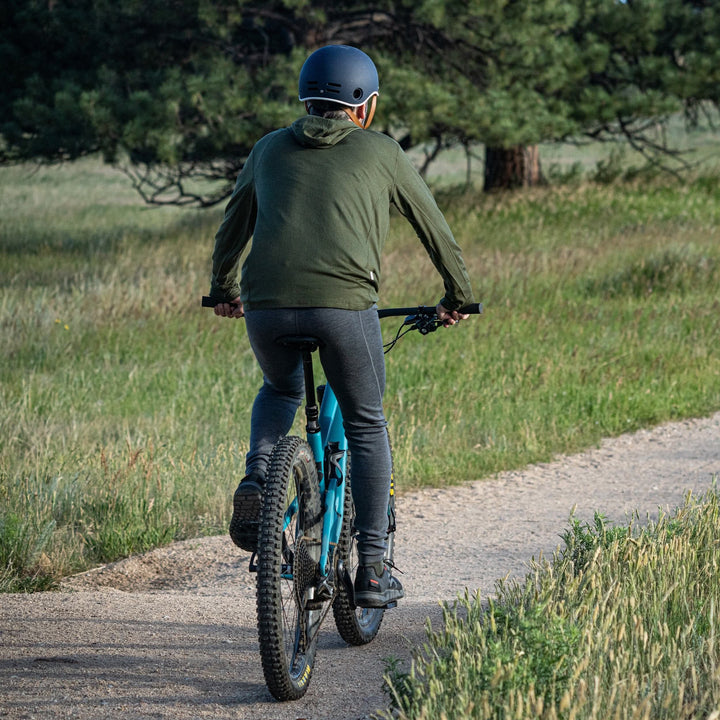 Back action view of a man riding a blue mountain bike along a gravel trail in charcoal gray midweight merino wool flyless base-layer bottoms, an olive hooded shirt, black shoes, and a navy helmet 
