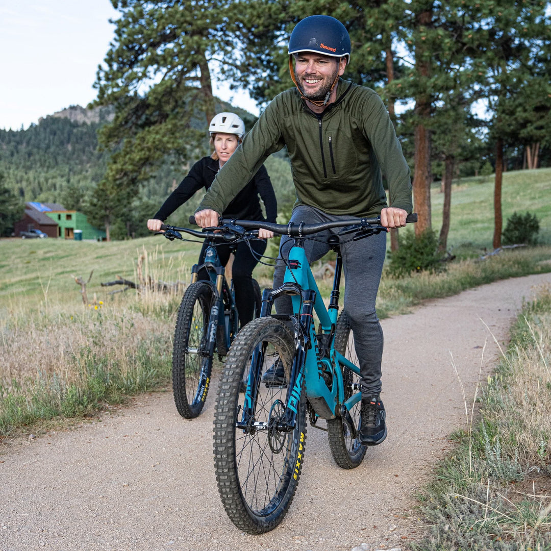 Front action view of a smiling man mountain biking on a dirt trail in gray midweight merino wool flyless base-layer bottoms, an olive quarter-zip top, and navy helmet, with a second helmeted rider following behind