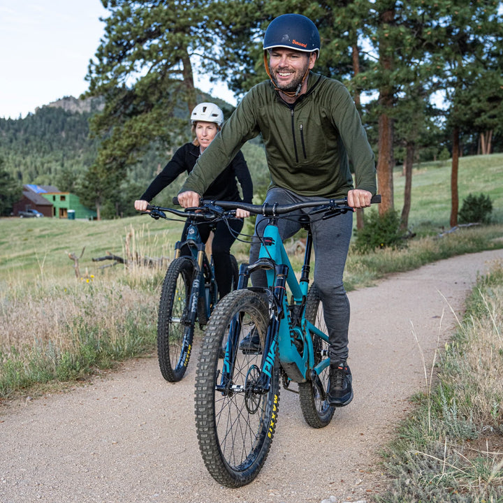 Front action view of a smiling man mountain biking on a dirt trail in gray midweight merino wool flyless base-layer bottoms, an olive quarter-zip top, and navy helmet, with a second helmeted rider following behind