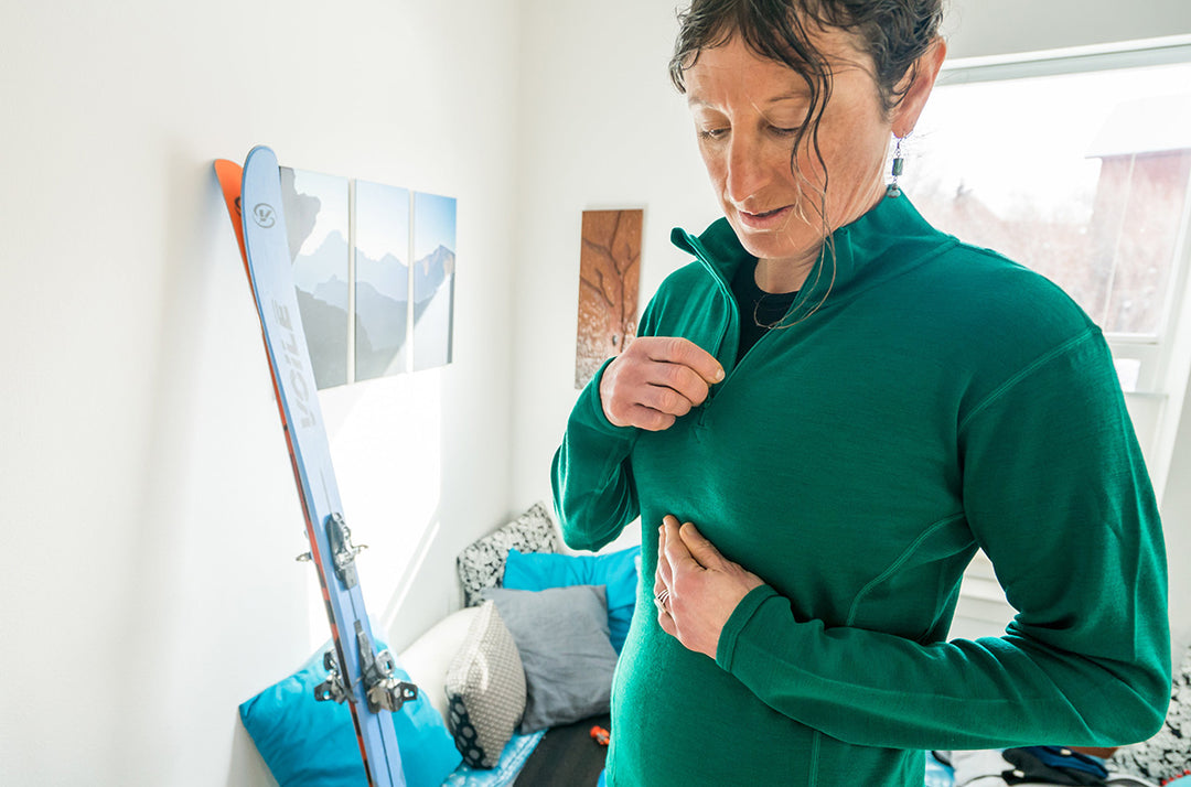 Front three-quarter view of a woman preparing to ski while zipping a midweight merino wool quarter-zip top, showing the stand collar, chest zipper, fitted sleeves, and layered ski clothing