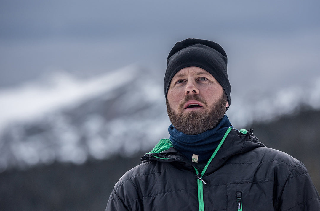 Close-up front view of a bearded man wearing a black insulated Thermerino wool jacket with bright green zipper trim, a navy neck gaiter, and a black knit beanie against snowy mountain peaks