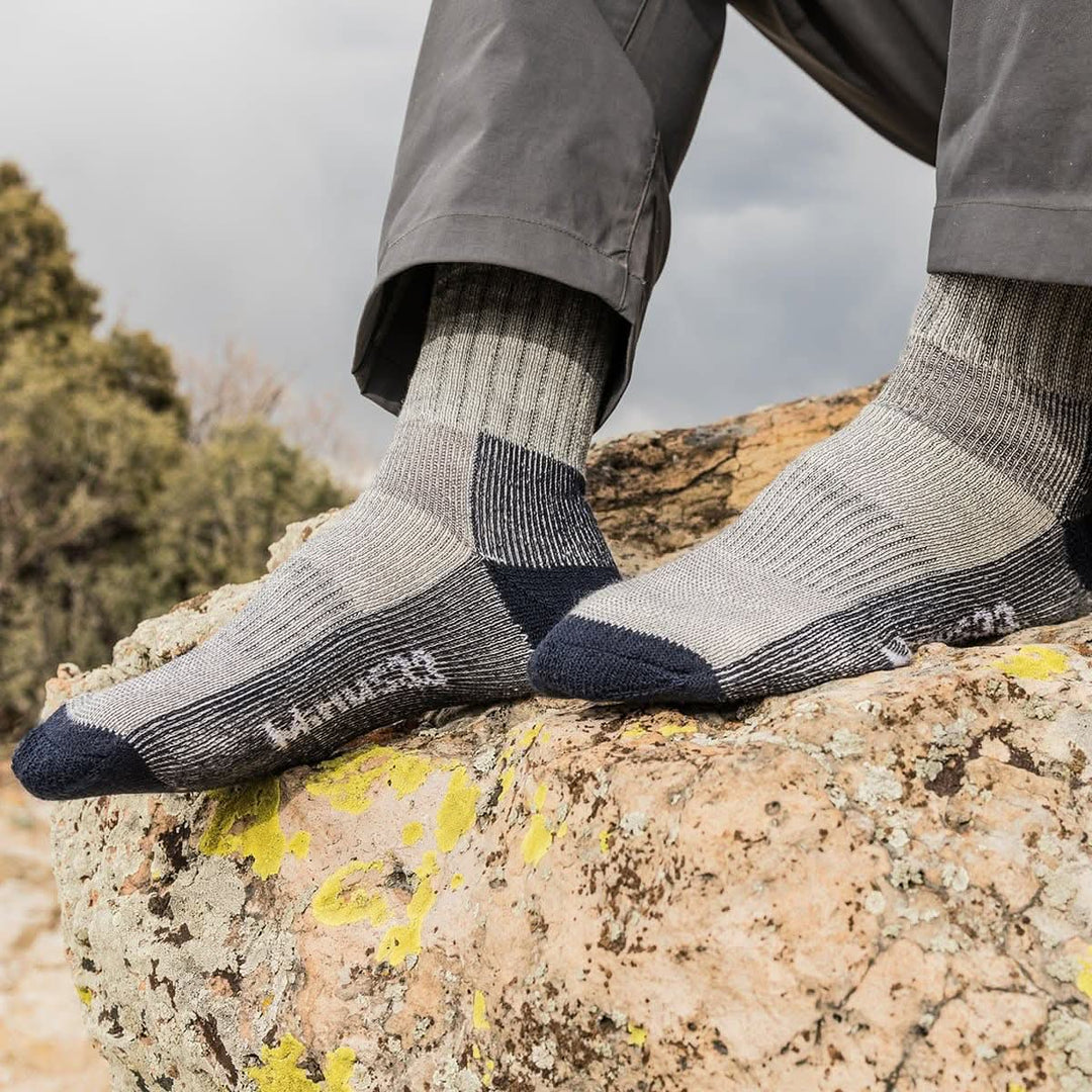 Close-up side view of both feet wearing denim heather and gray merino wool day-hiker socks on a lichen-covered rock, showing navy reinforced toes and heels, ribbed arch panels, and Minus33 logos below gray hiking pants 