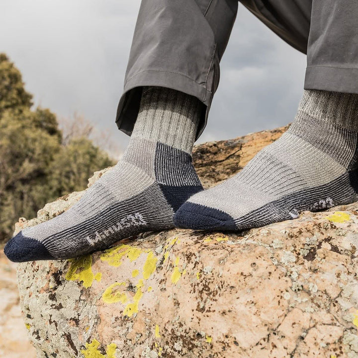 Close-up side view of both feet wearing denim heather and gray merino wool day-hiker socks on a lichen-covered rock, showing navy reinforced toes and heels, ribbed arch panels, and Minus33 logos below gray hiking pants 
