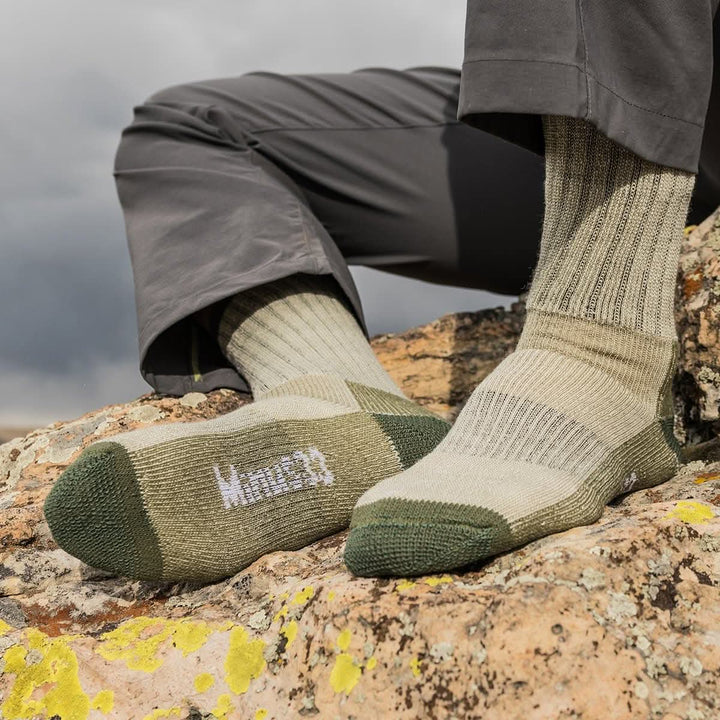 Close-up front and sole view of both feet wearing olive green merino wool day-hiker socks on a lichen-covered rock beneath gray hiking pants, showing dark green reinforced toes and heels, ribbed panels, and a white Minus33 sole logo 