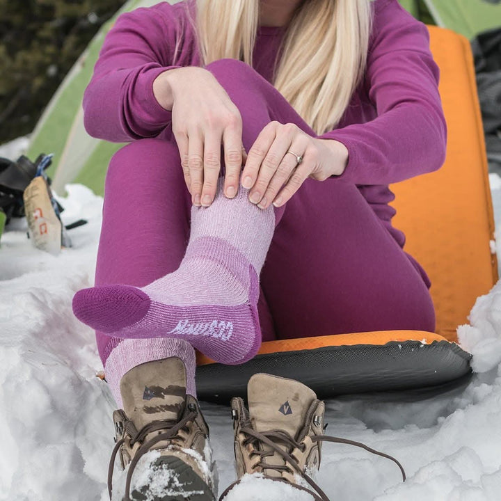 Front close-up view of a woman seated on an orange pad in snow wearing a radiant violet merino wool base-layer set while pulling up a lilac and purple day-hiker sock beside unlaced brown hiking boots 