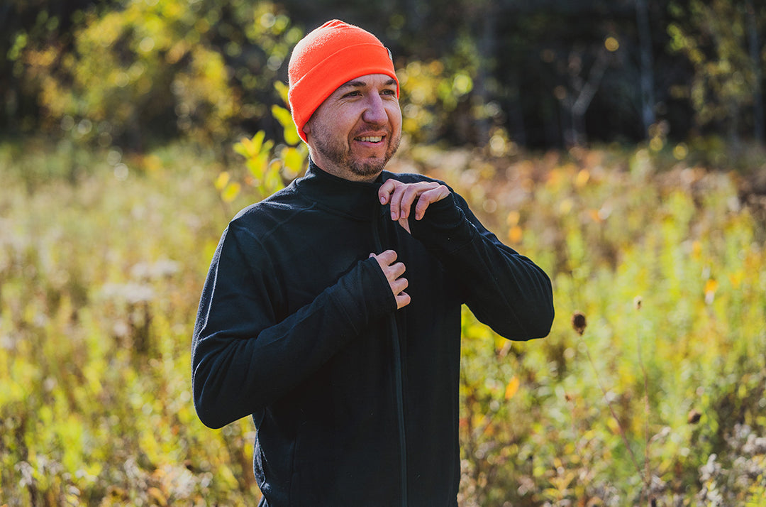 Waist-up three-quarter front view of a smiling man in a blaze-orange merino wool beanie adjusting the zipper of a black wool Wilderness midlayer in a sunlit grassy field