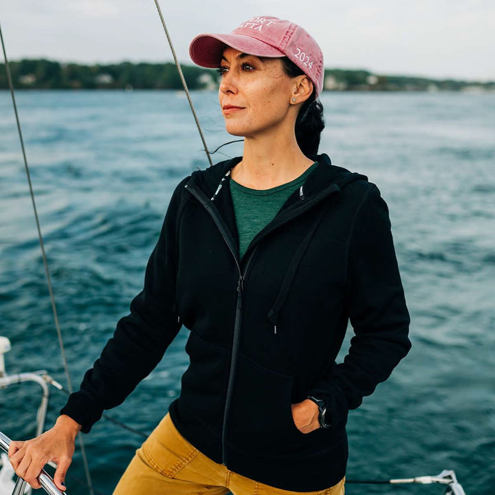 Woman on a sailboat wearing a black wool fleece jacket hoodie and pink cap against a water background. 