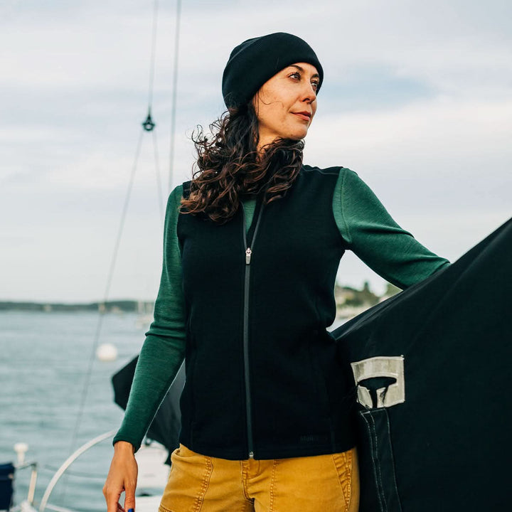 Woman wearing a green and black merino wool sweater vest jacket with a sailboat in the background 